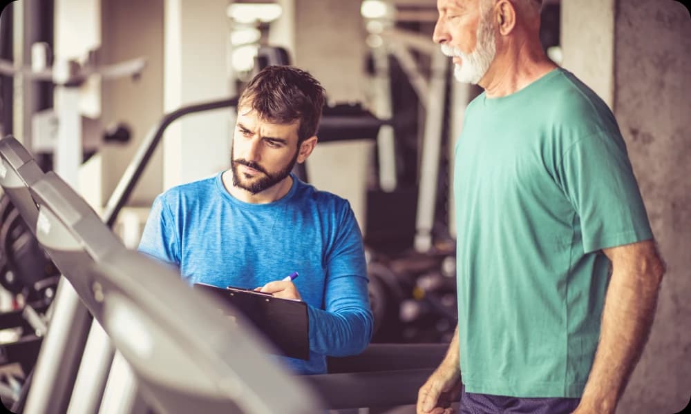 senior citizen on a treadmill with a personal trainer alongside him noting down his progress